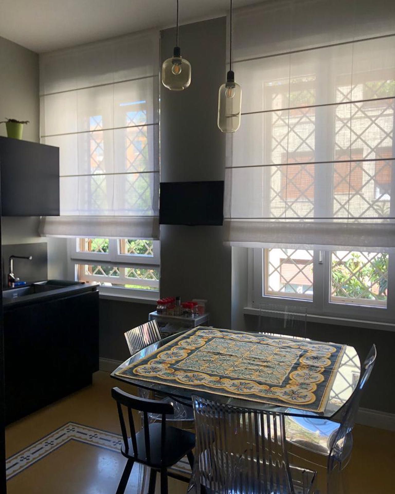 Zona pranzo con tavolo in vetro, quattro sedie, due lampade a sospensione e due finestre con tende trasparenti.