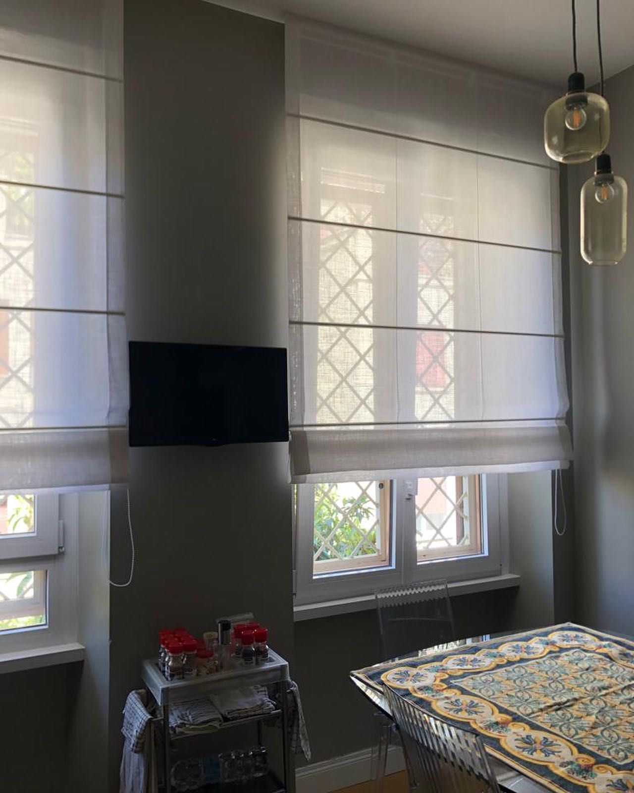 Stanza piena di luce con tende romane bianche alle finestre, TV a parete, tavolino e lampade sospese.