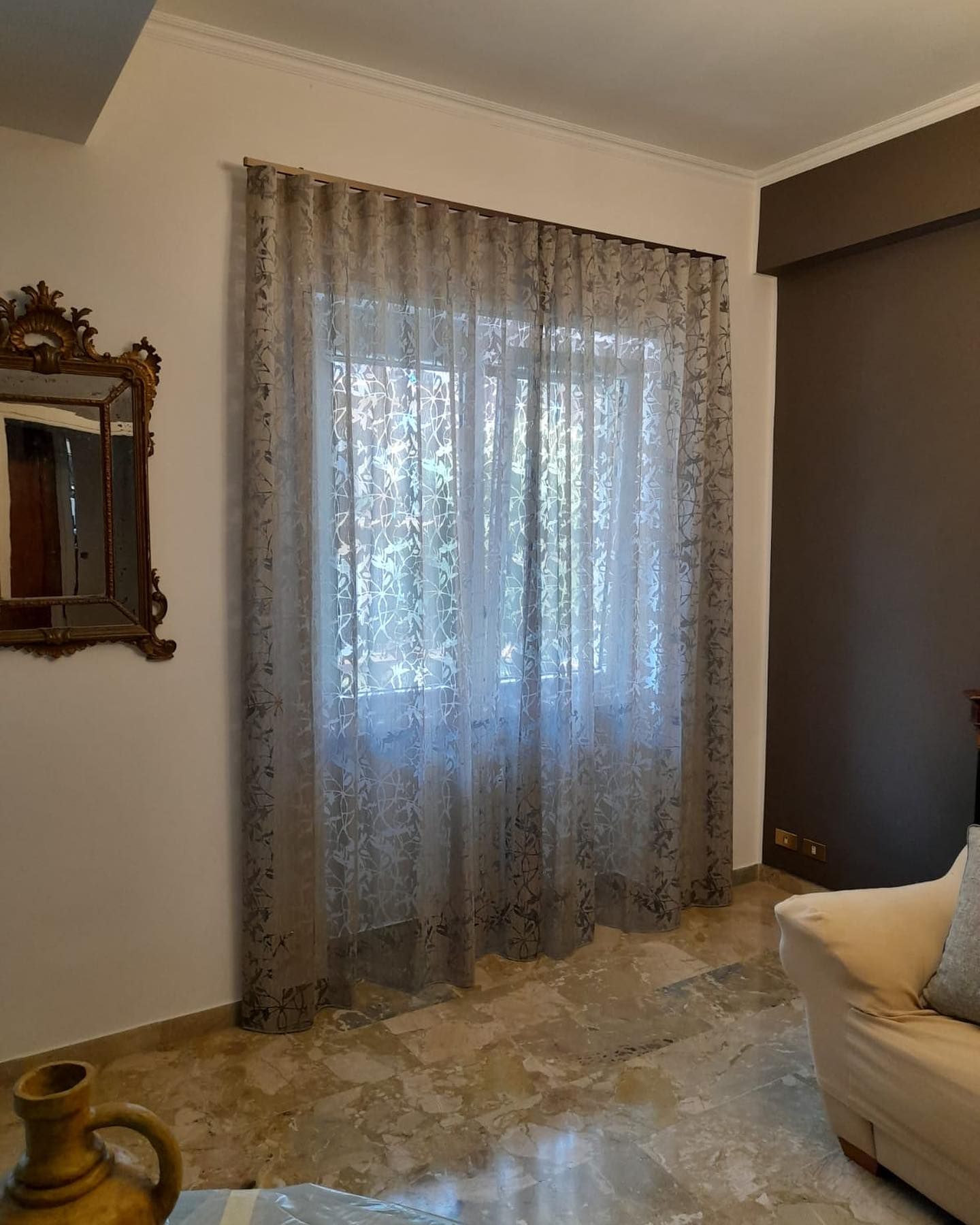 Finestra con tende trasparenti a fantasia, specchio sulla parete e pavimento in marmo in una stanza.