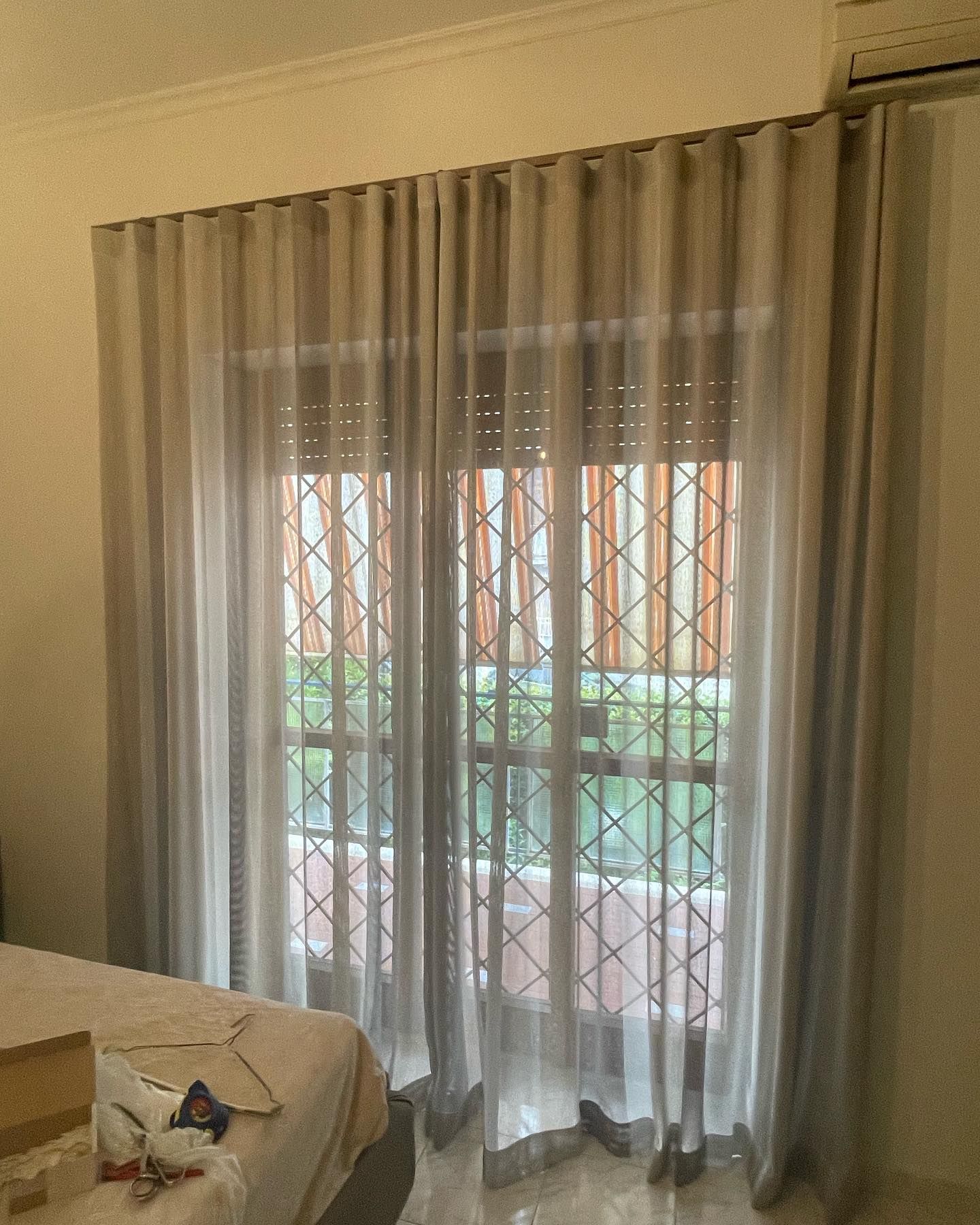 Tende beige che incorniciano una finestra con zanzariera; letto visibile sulla sinistra.