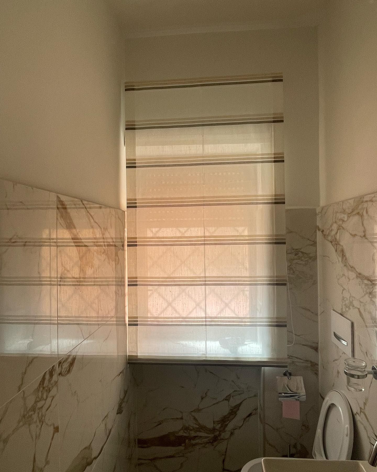 Finestra del bagno con tenda romana sopra le piastrelle e un WC.