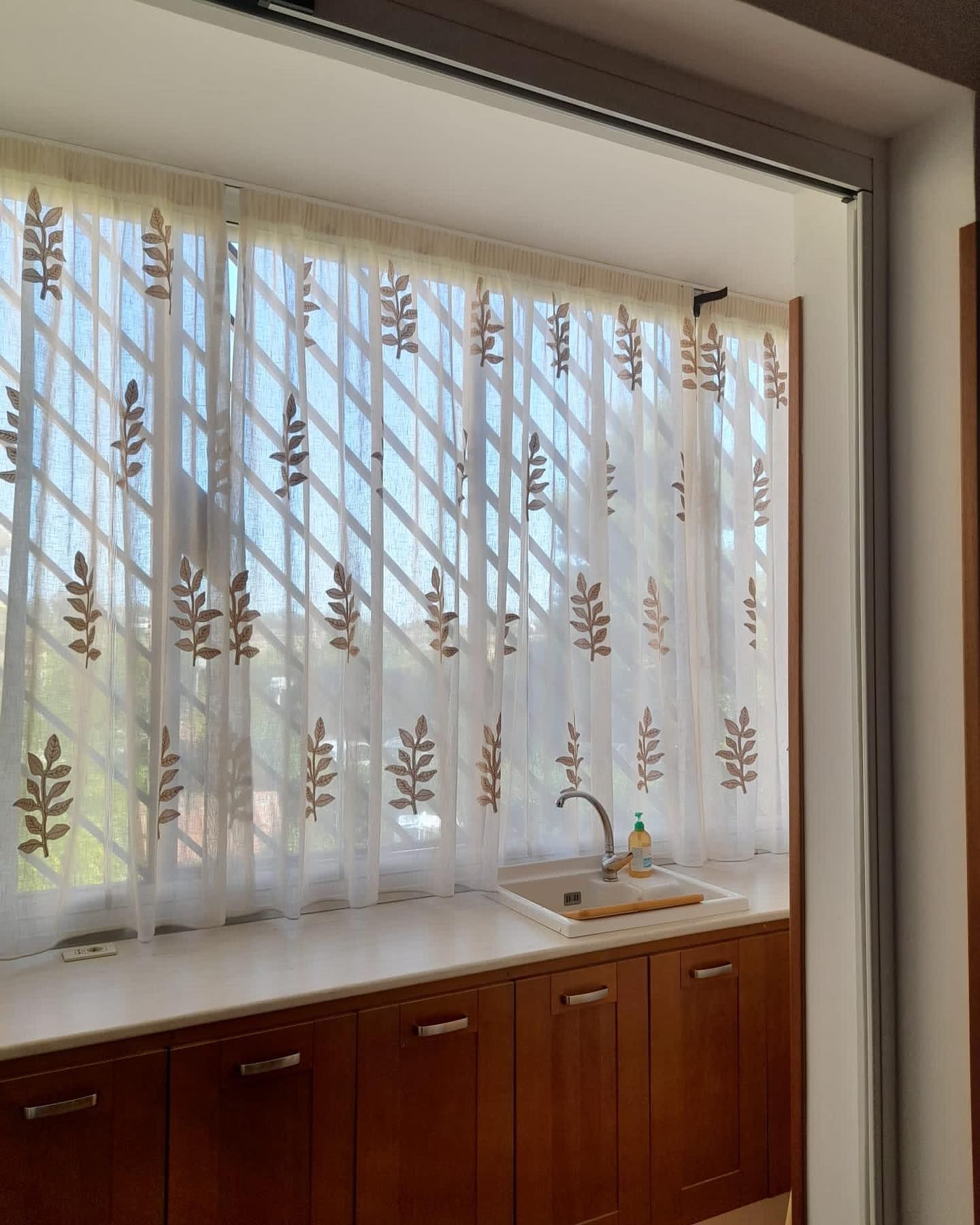 Finestra della cucina con tende trasparenti con motivi a foglie sopra un piano di lavoro con lavello e mobili in legno.