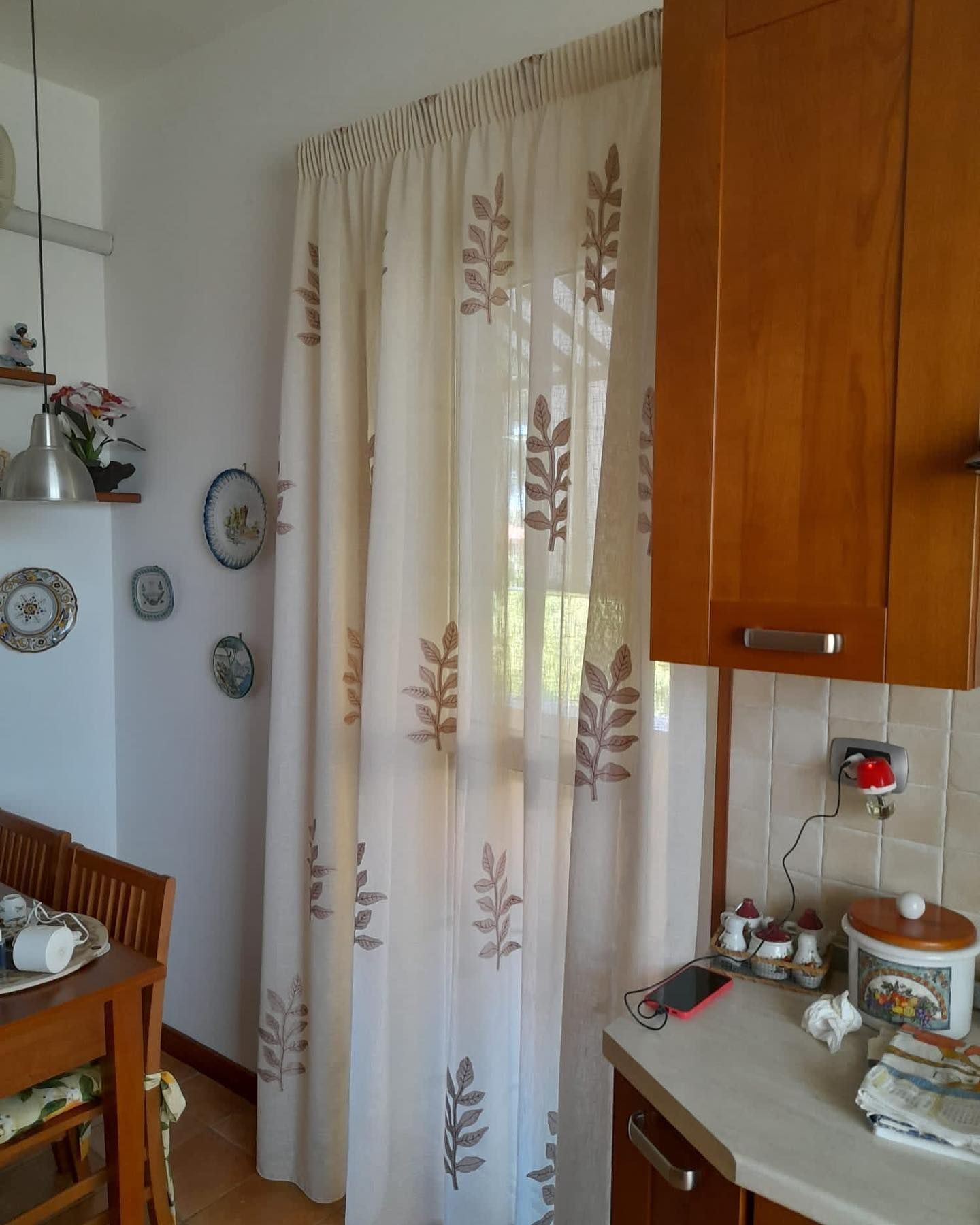 Cucina con tende dai colori chiari con motivi a foglie marroni, finestra e mobili in legno.