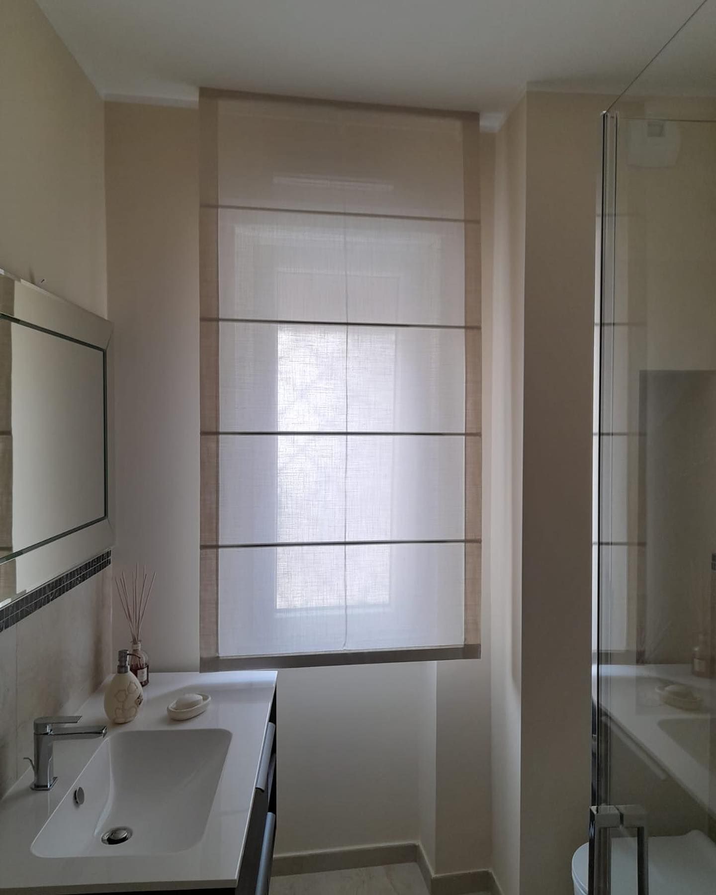 Bagno con tenda romana beige sopra una finestra, sopra un lavandino e uno specchio.