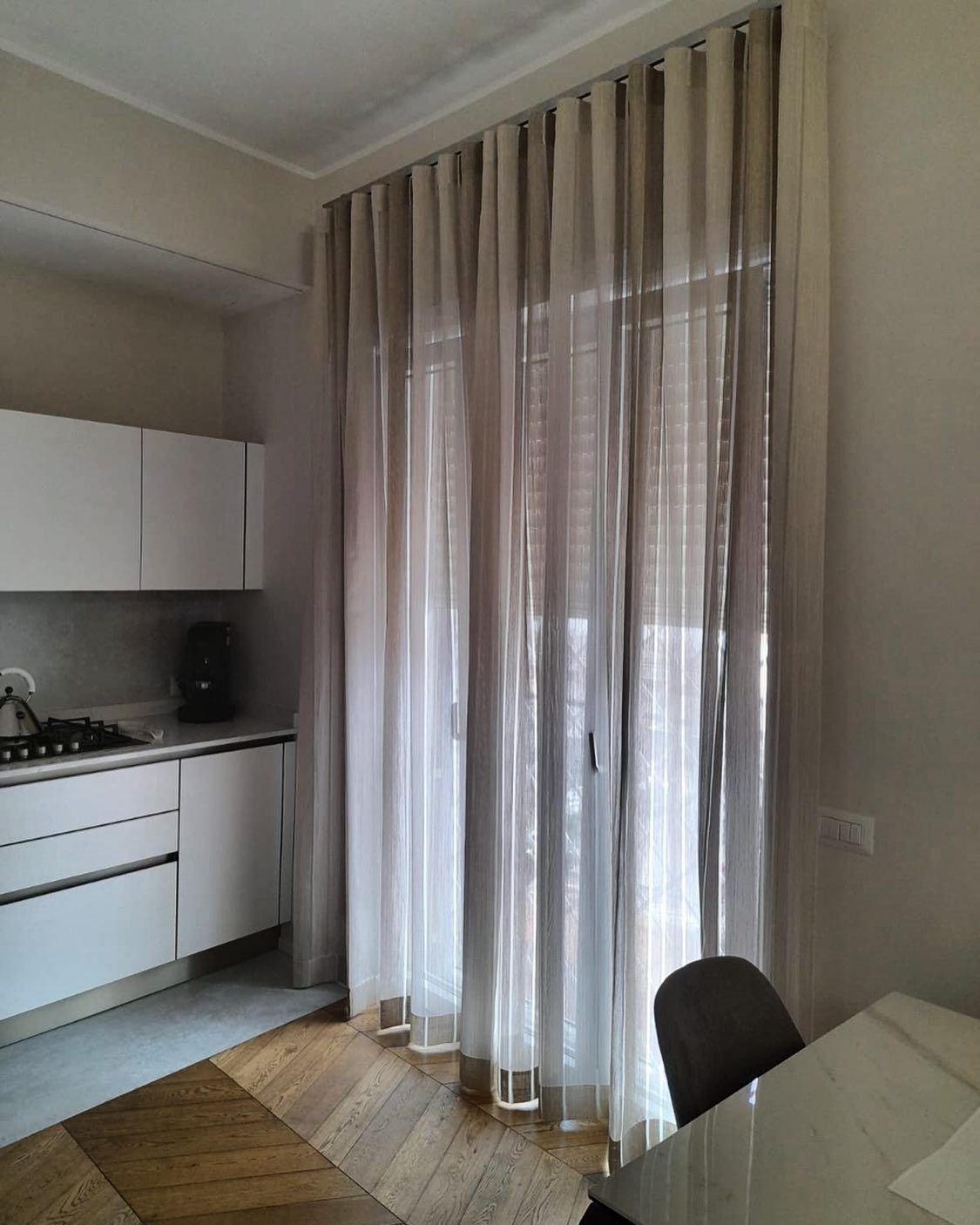 Cucina con mobili bianchi, tende beige trasparenti e pavimento in legno. Un tavolo con sedia davanti.