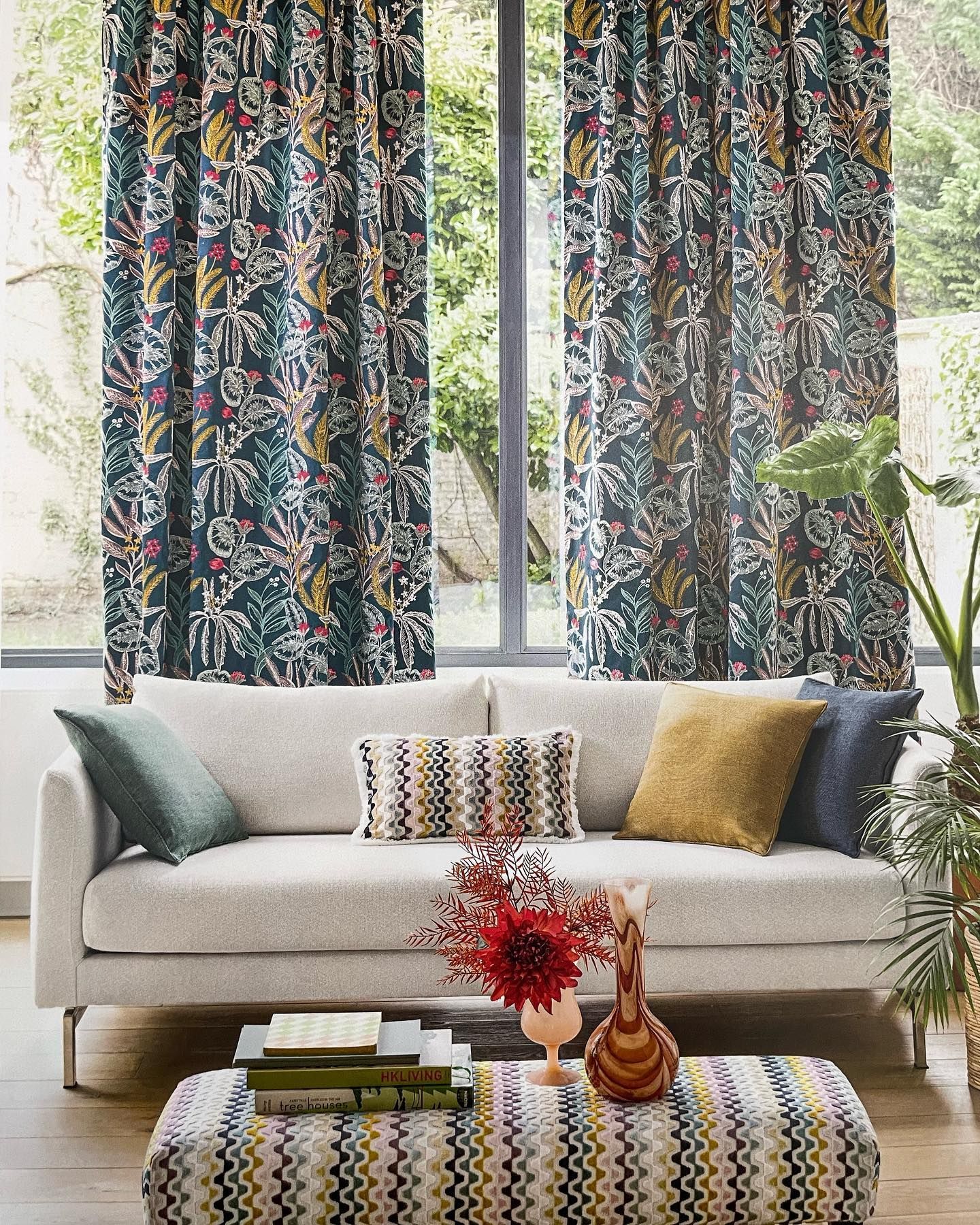 Soggiorno con tende stampate, divano color crema con cuscini colorati e pouf fantasia.