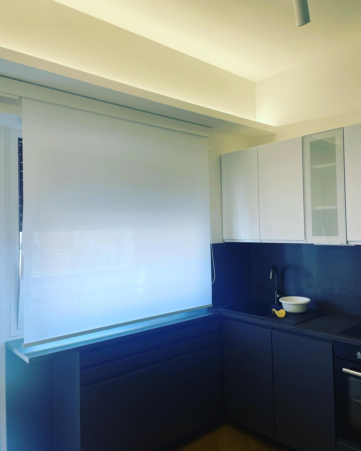 Cucina con mobili bianchi, ripiani e mobili blu scuro e una tenda a rullo bianca sopra la finestra.