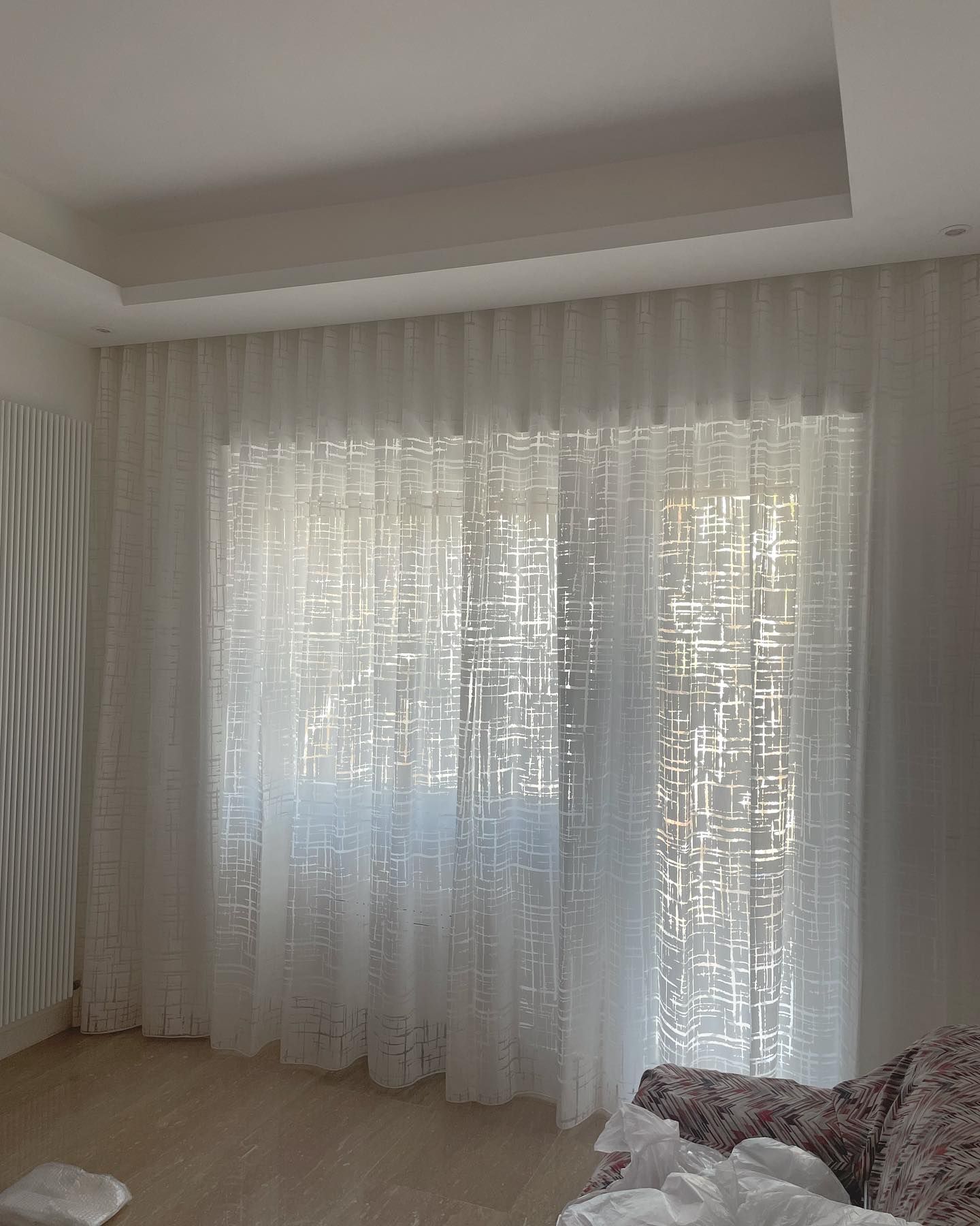 Tende bianche trasparenti davanti a una finestra, con un motivo strutturato; sono visibili il soffitto e le pareti bianche.