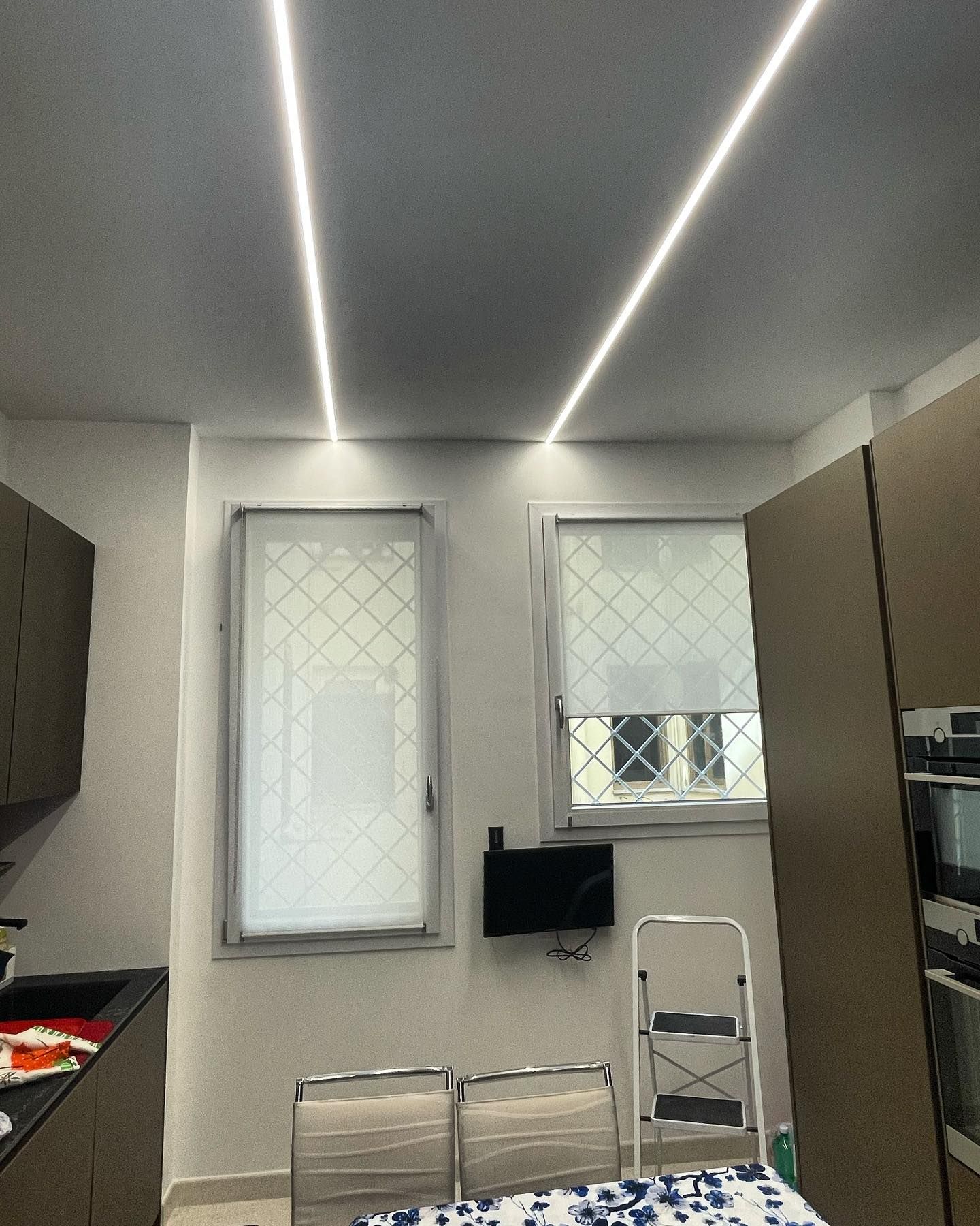 Luci lineari luminose sul soffitto in una stanza con due finestre e una piccola TV.