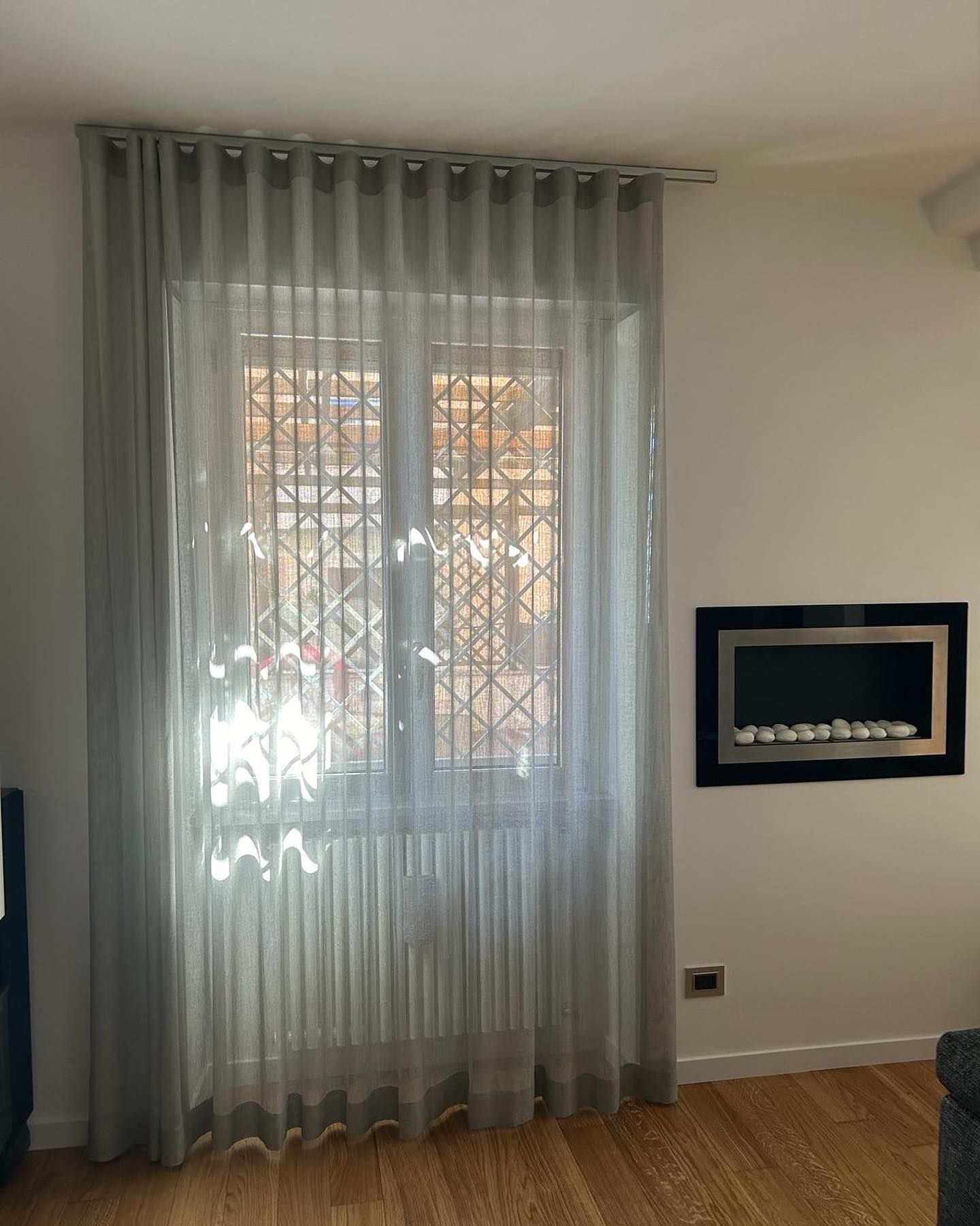 Tende grigie trasparenti coprono una finestra. Un camino è incastonato nella parete bianca sulla destra.