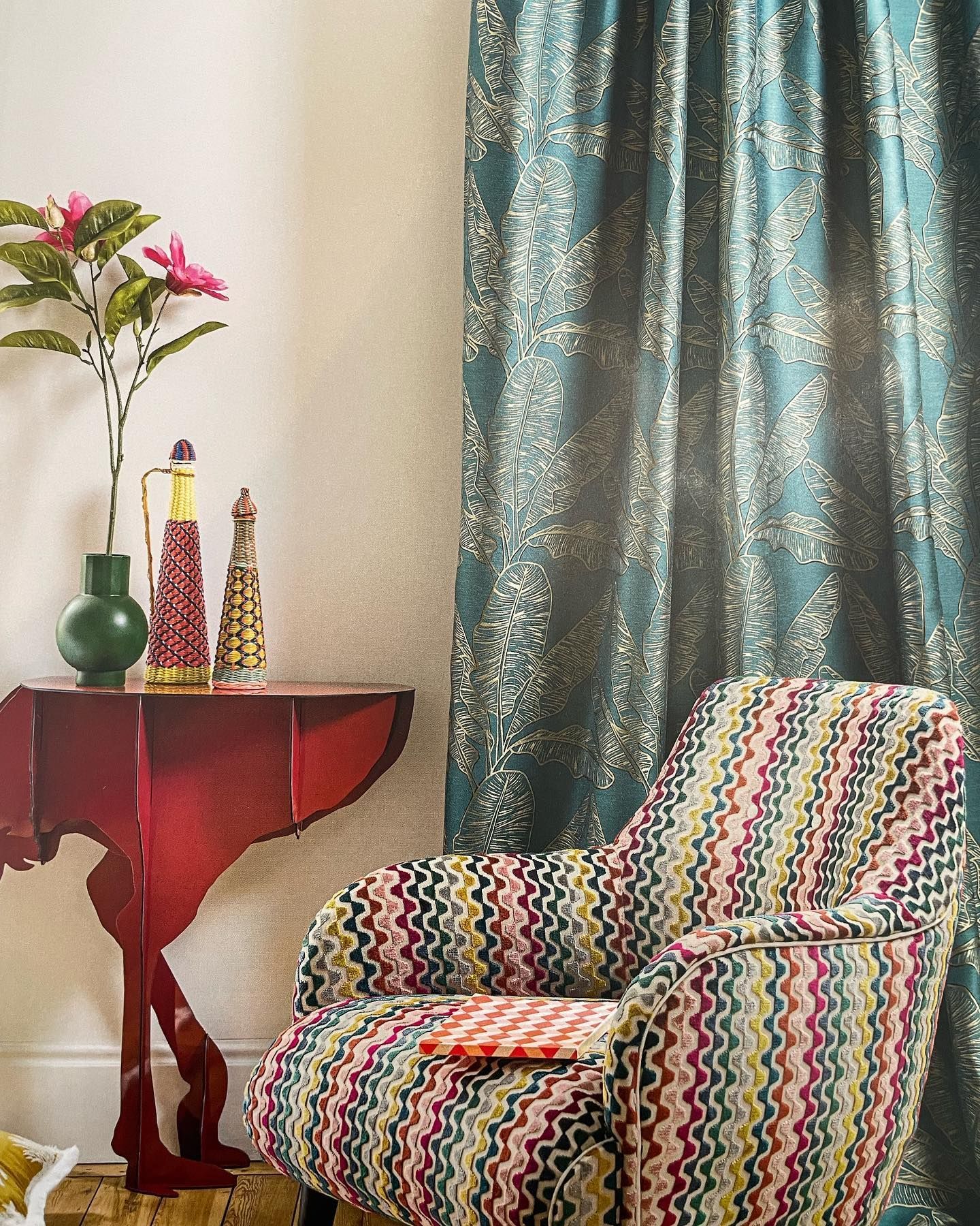 Tavolo rosso con fiori e statue, poltrona con motivi e tenda con motivi color verde acqua.