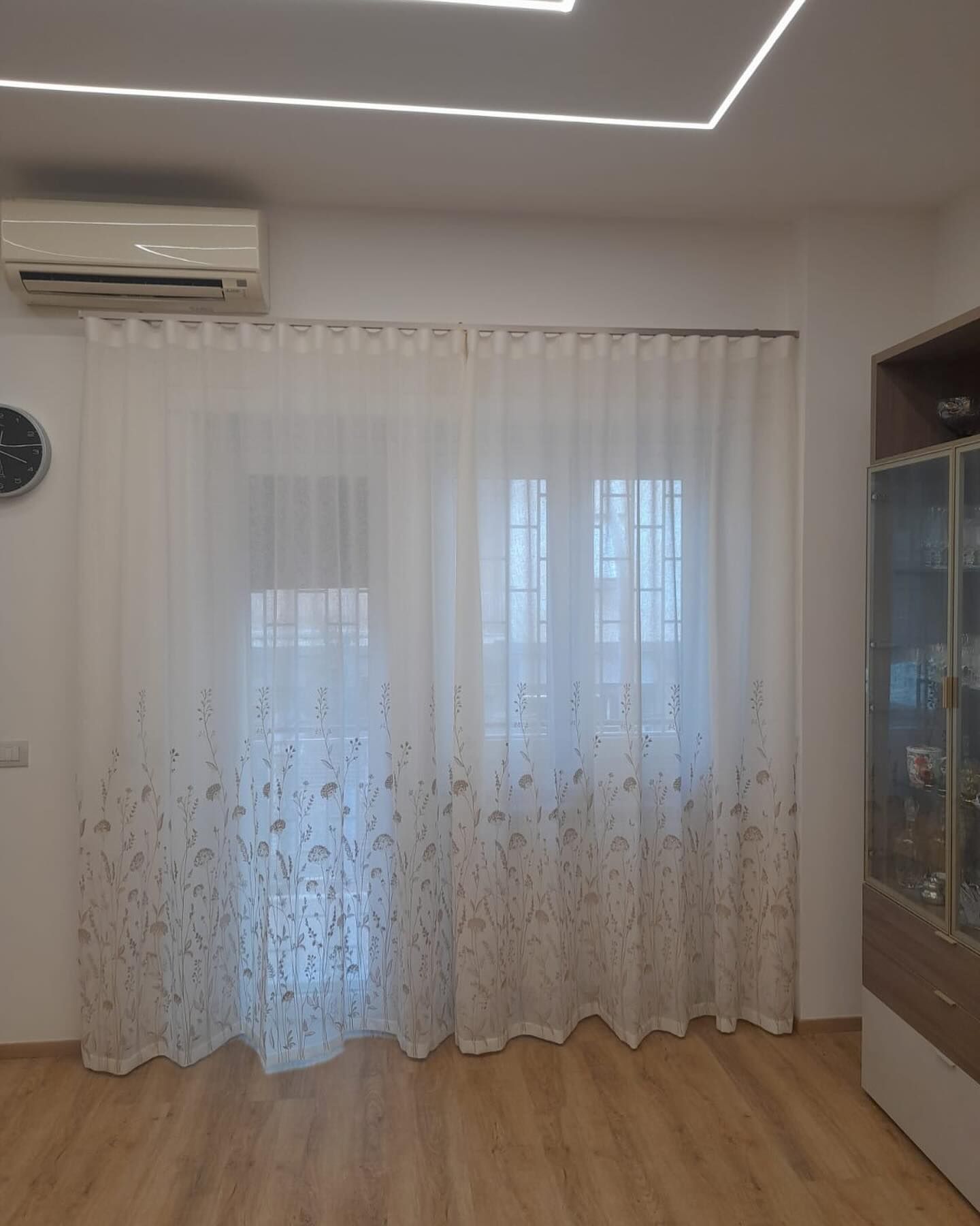 Tende bianche trasparenti che coprono una finestra. Unità di aria condizionata sopra le tende. Pavimento e mobili in legno.