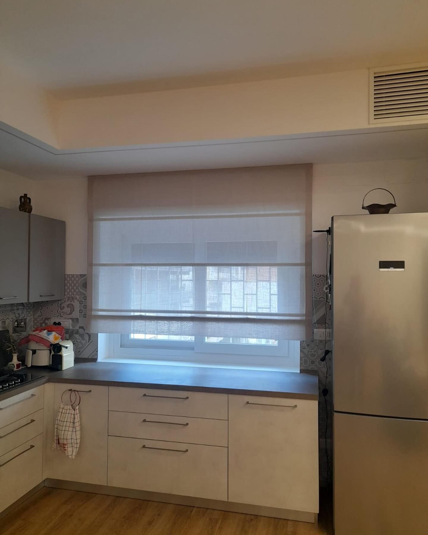 Cucina con mobili bianchi, piano di lavoro grigio, finestra con tende romane di colore chiaro