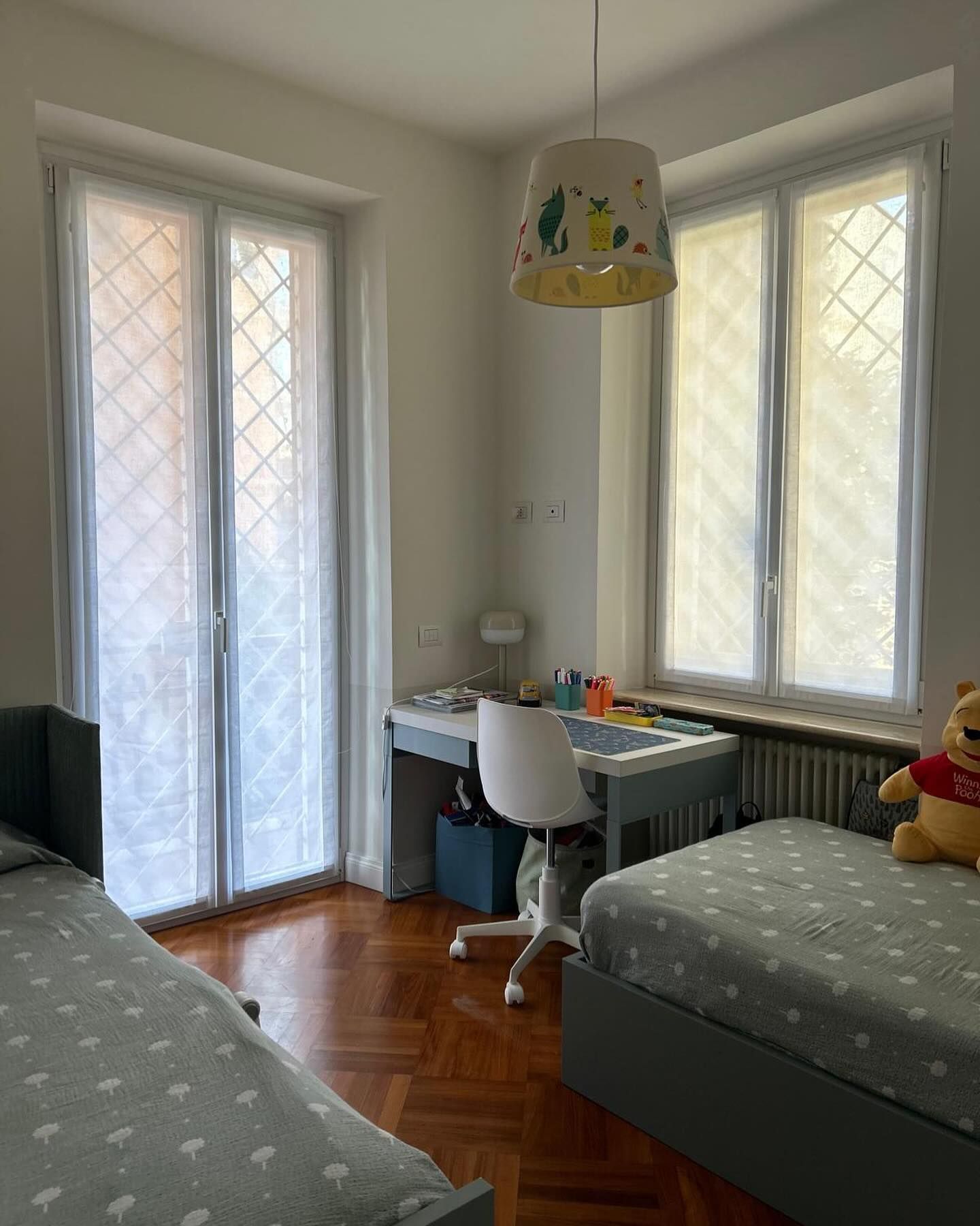 Camera da letto con due letti singoli, una scrivania e finestre con tende trasparenti.