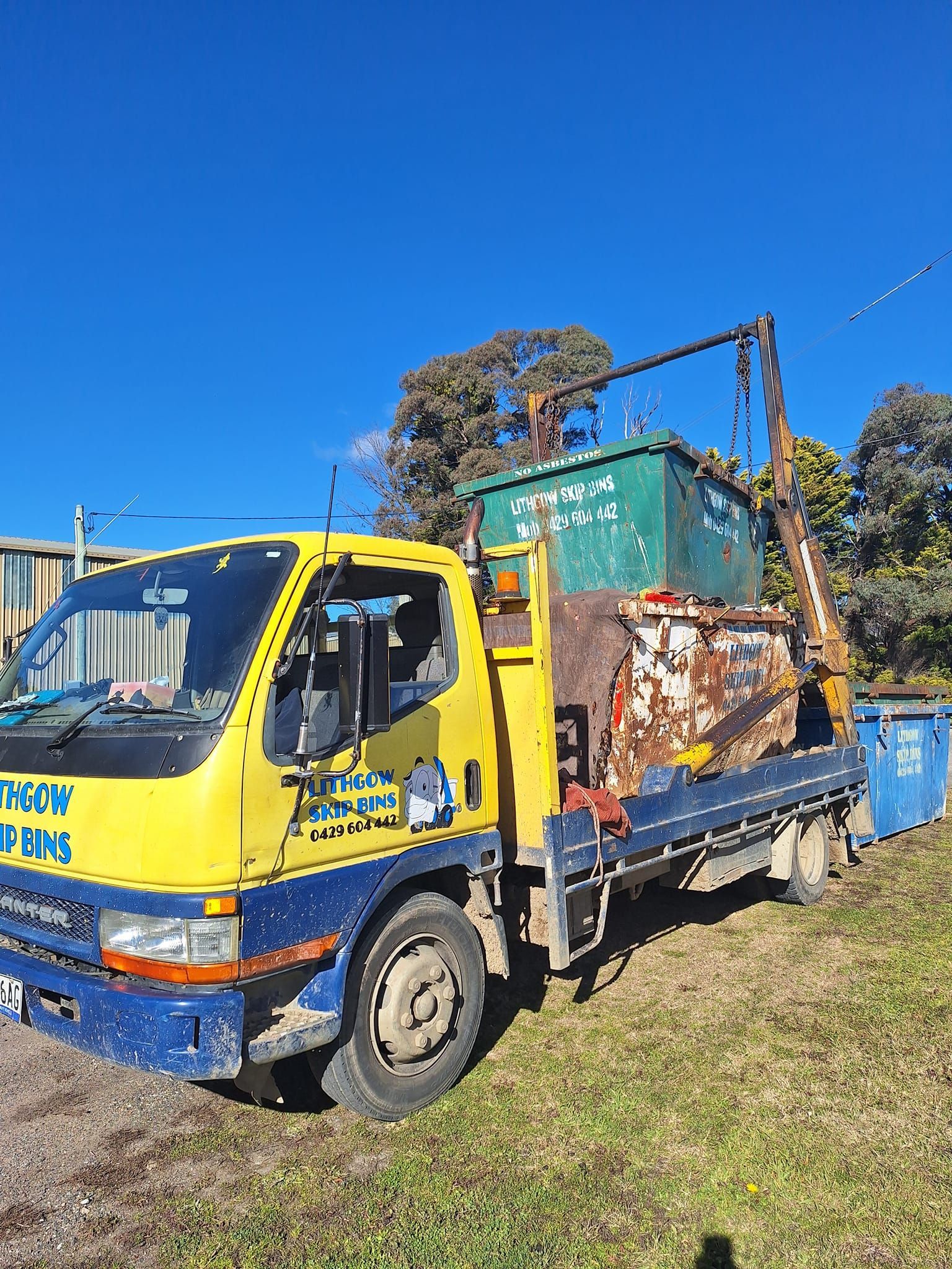 Green dumpster sits on grass near trees.
