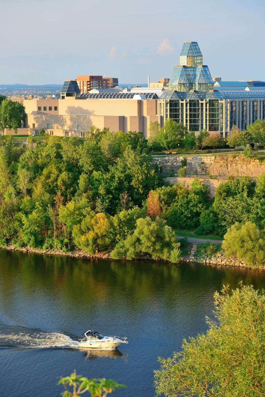 Riverfront cityscape with a large modern building behind green trees and a waterfall in the foreground