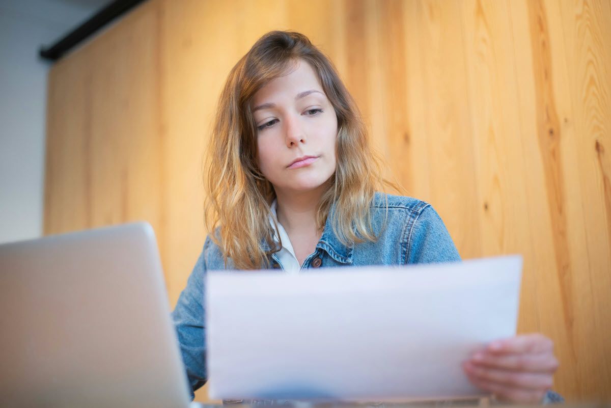 Woman in denim jacket looks at papers, laptop visible, wooden backdrop.