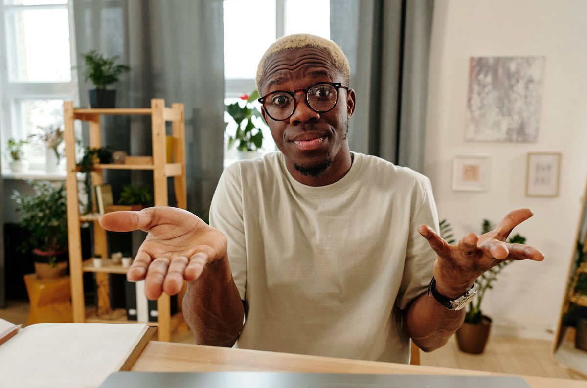 Man in glasses shrugging at a desk in a bright home office, with shelves and plants in the background