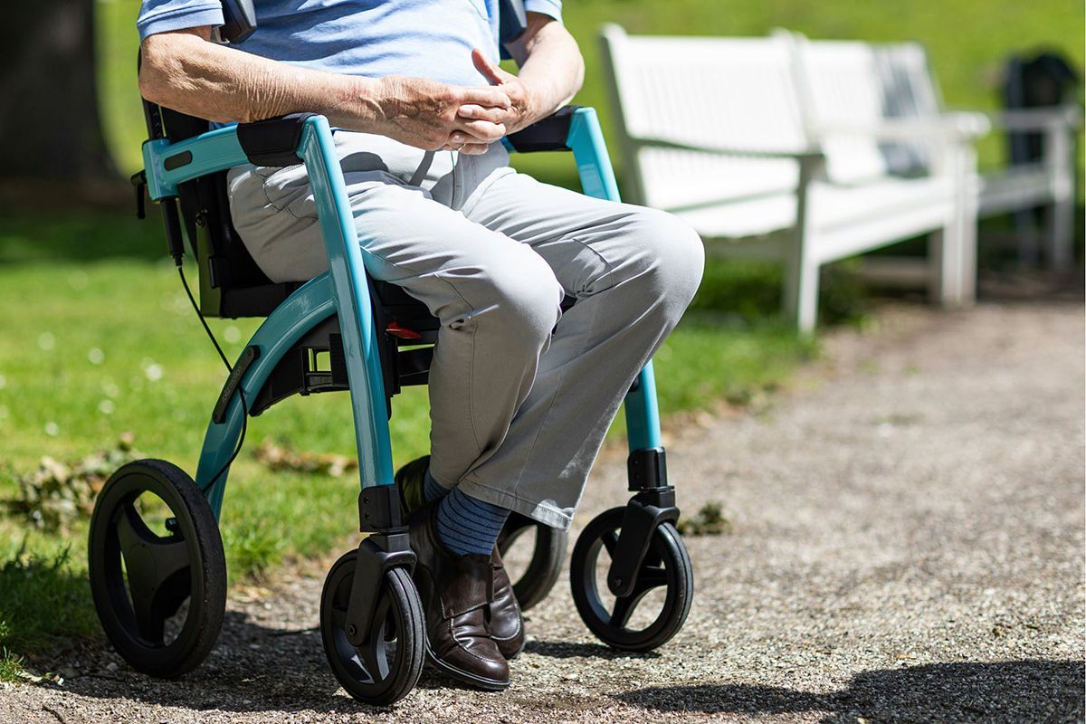 Person in light-colored pants sitting in teal walker on a paved path in a park with a white bench in the background.