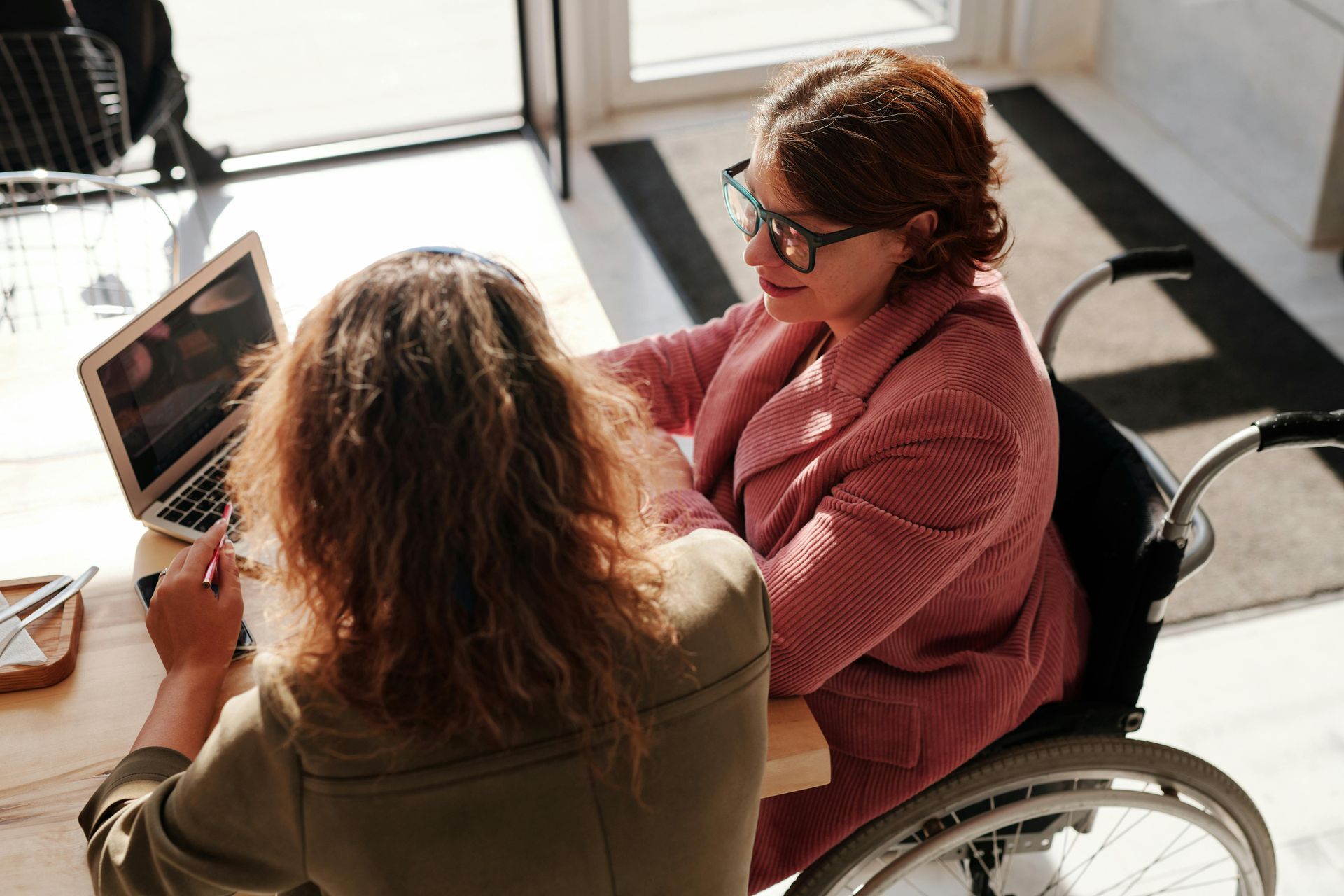 A woman in a wheelchair is sitting at a table with another woman using a laptop computer.