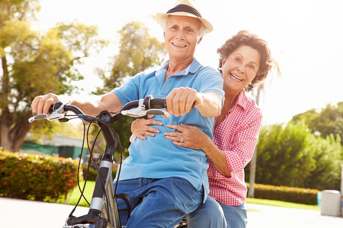 Elderly couple riding a bicycle, smiling outdoors, woman embracing man.