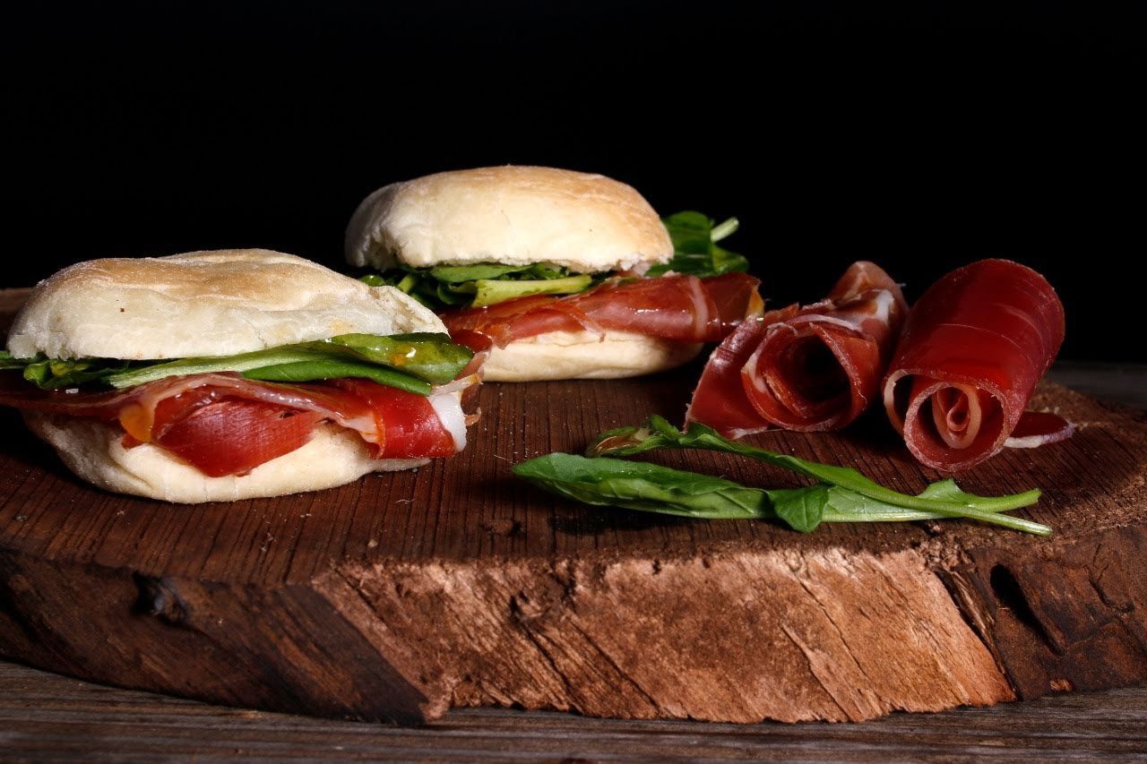 Dos bocadillos con verduras y tomates, acompañados de embutidos enrollados sobre una tabla de madera.