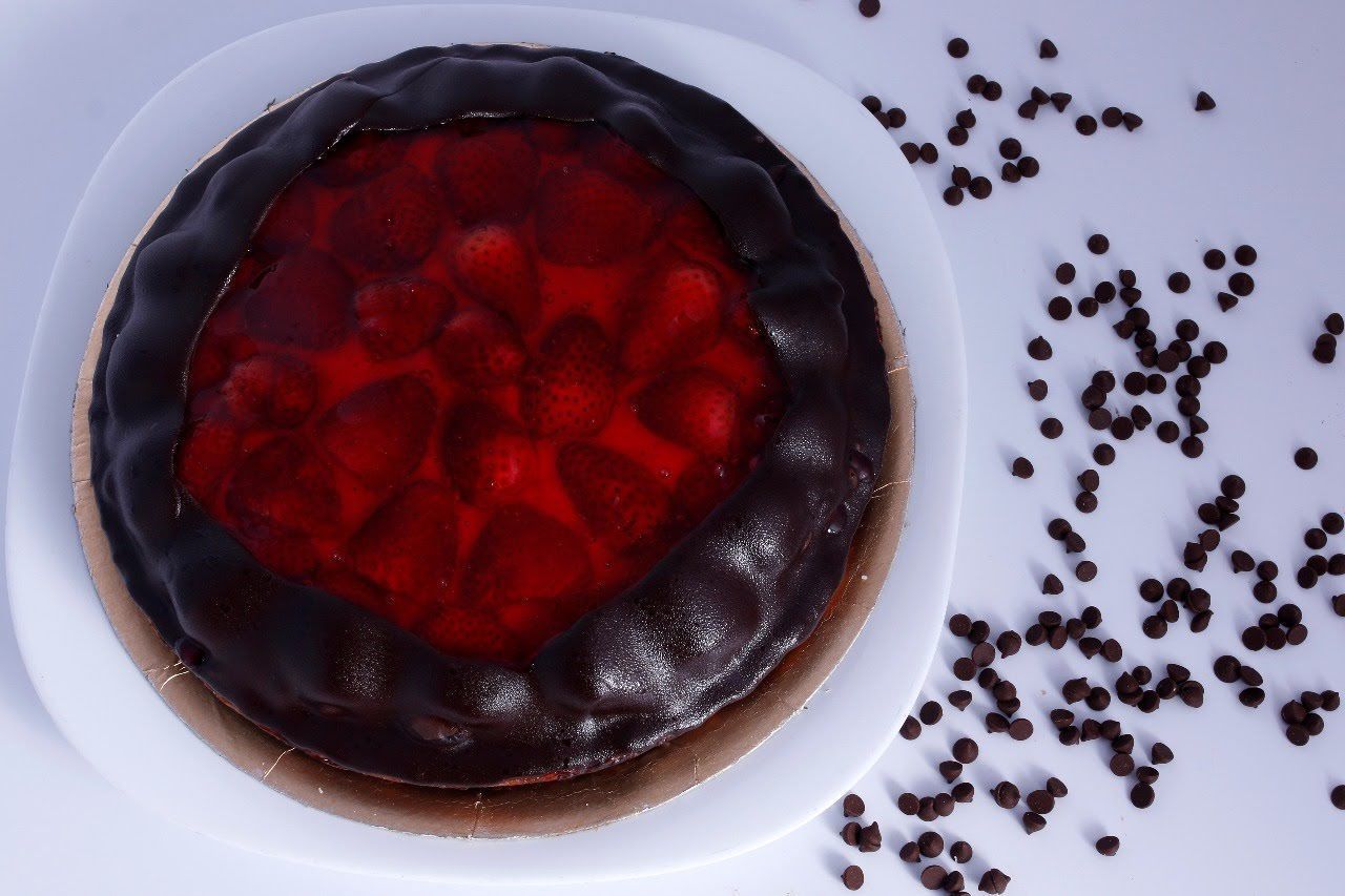 Tarta de chocolate con una brillante cobertura de frutos rojos sobre un plato blanco, con granos de café esparcidos cerca.