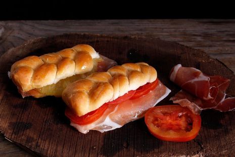 Sándwich de jamón curado y tomate en una tabla de madera rústica, con lonchas de prosciutto al lado.