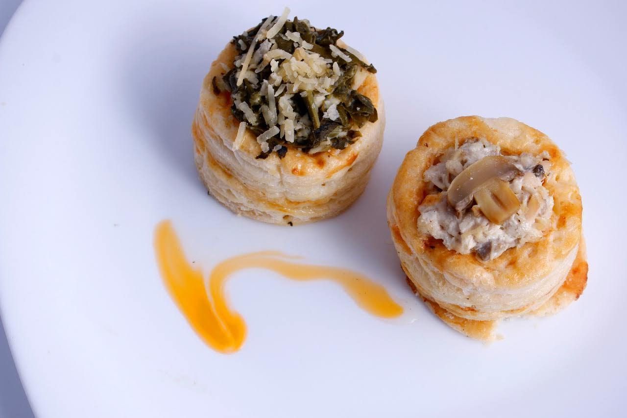 Dos bocaditos salados de hojaldre en un plato blanco, uno cubierto con hierbas y el otro con champiñones, junto a una salsa.
