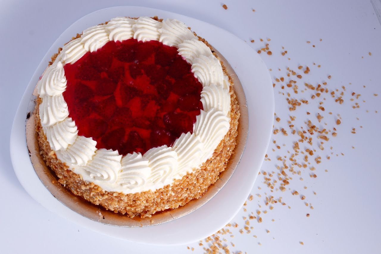 Pastel redondo con centro de gelatina roja, borde de crema blanca y cobertura de migas sobre un plato blanco.