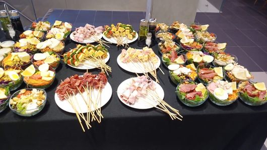 Mesa de buffet con muchos platos pequeños y coloridos para aperitivos y brochetas sobre un mantel negro.