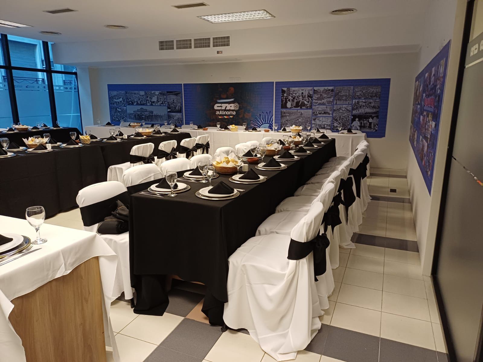 Comedor preparado para un banquete formal con largas mesas negras, sillas blancas y vajilla.