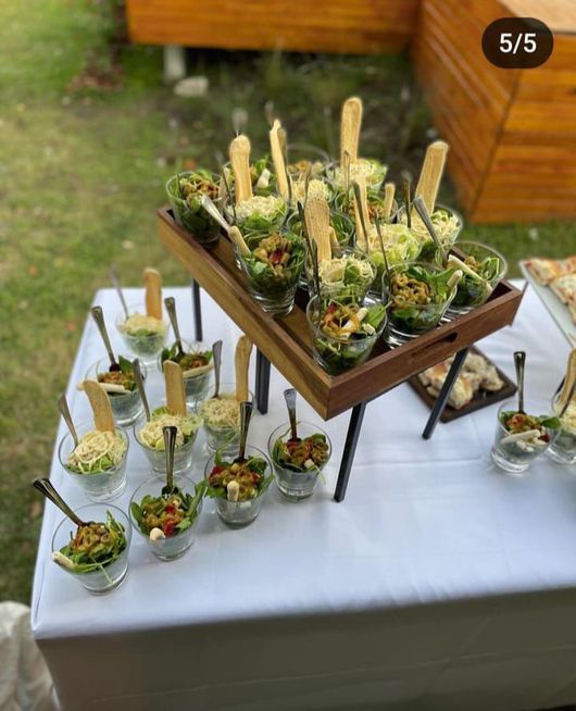 Buffet de postres sobre una mesa blanca con tazas pequeñas y cucharas de madera, dispuesto al aire libre sobre una bandeja.