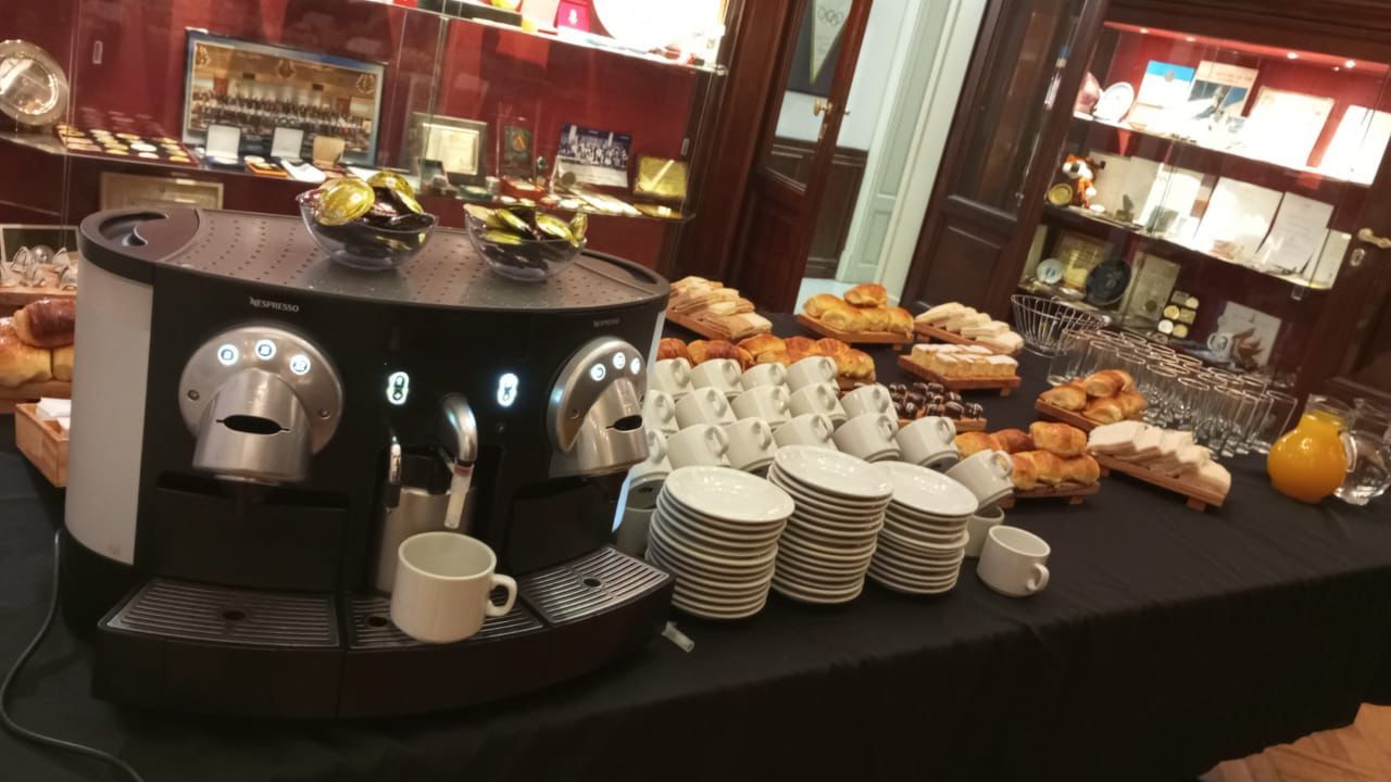 Mesa de buffet con café y pasteles, con tazas, platos, pasteles apilados y un dispensador de bebidas.