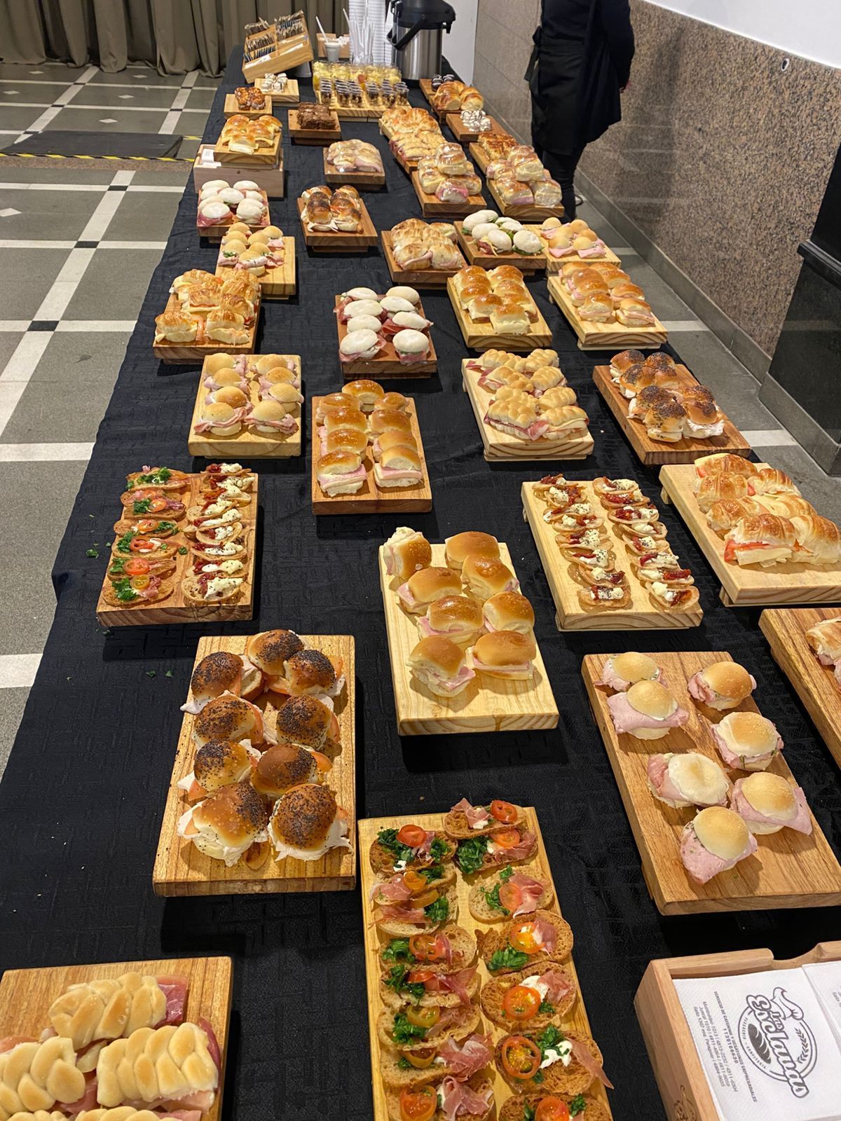Una larga mesa con una variedad de pasteles y postres expuestos en bandejas al aire libre, con dos personas al fondo.