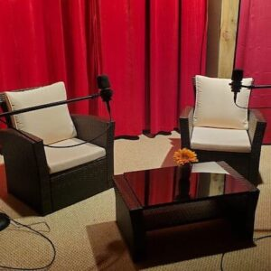 Two chairs with white cushions and microphones face a small black table against a backdrop of red curtains.