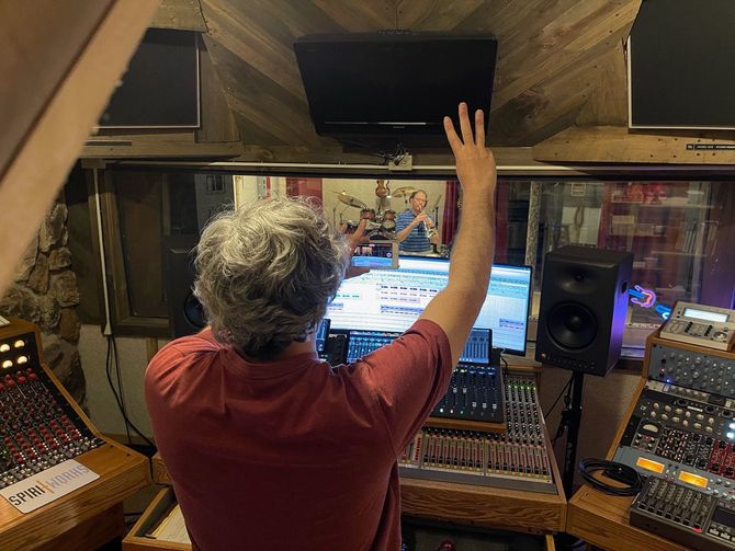 A person in a red shirt faces a studio control board with multiple monitors and audio equipment, reaching toward a screen.