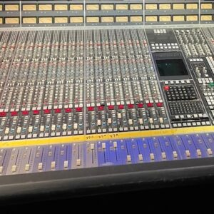 A large, professional audio mixing console with numerous knobs, sliders, and analog VU meters in a studio setting.
