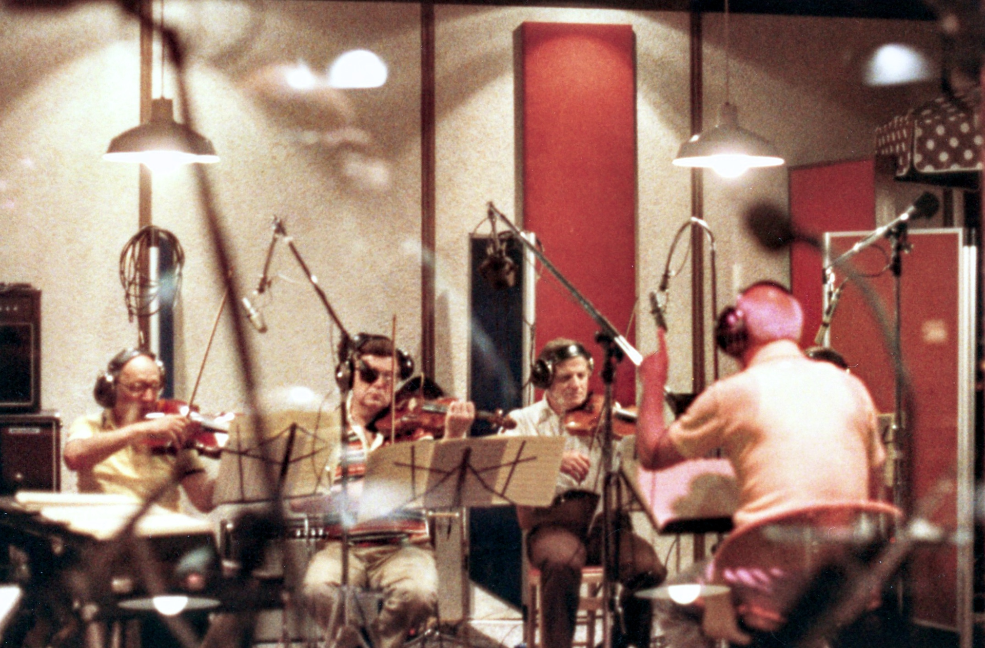 Four musicians with headsets playing violins in a sound-dampened recording studio with red acoustic panels.