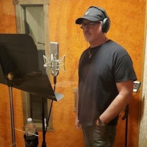 A person wearing headphones and a cap stands in a recording booth, facing a microphone and music stand with a water bottle.