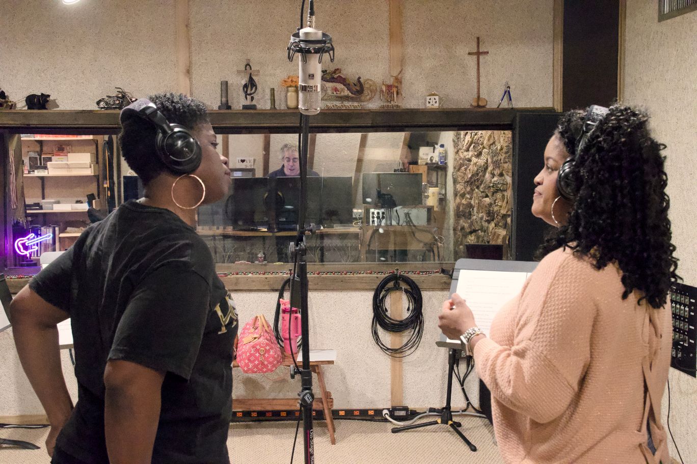 Two people in a recording studio wearing headphones stand before a microphone, looking at a script together.