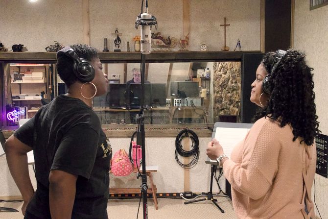 Two people in a recording studio wearing headphones stand before a microphone, looking at a script together.