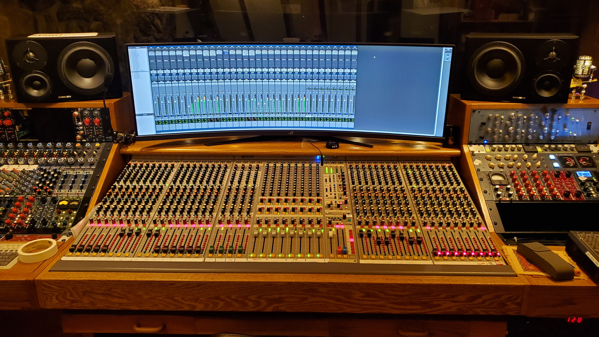 Professional recording studio featuring a large mixing console, dual studio monitors, and a widescreen computer display.