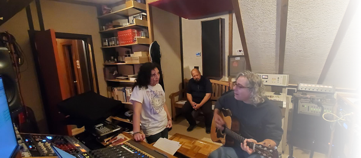 Three people in a home recording studio: one plays acoustic guitar while the others observe near a mixing desk.