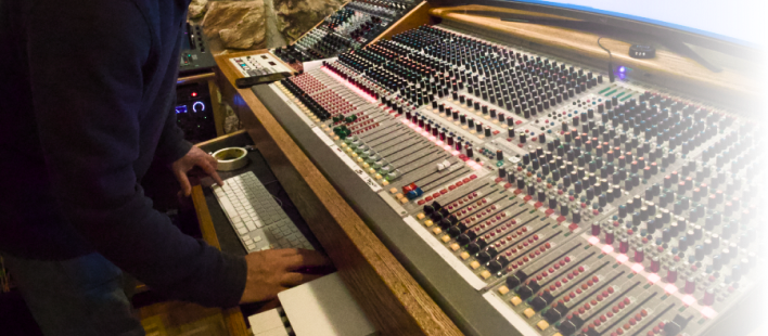 A person adjusting a keyboard in front of a large, complex audio mixing console in a recording studio.