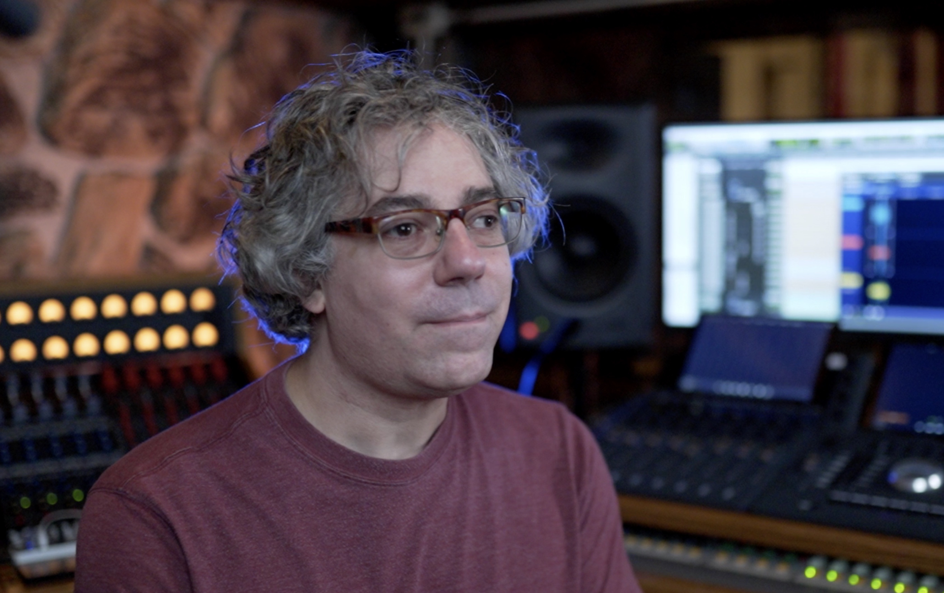 A person with curly hair and glasses in a recording studio, seated in front of audio mixing equipment and monitors.