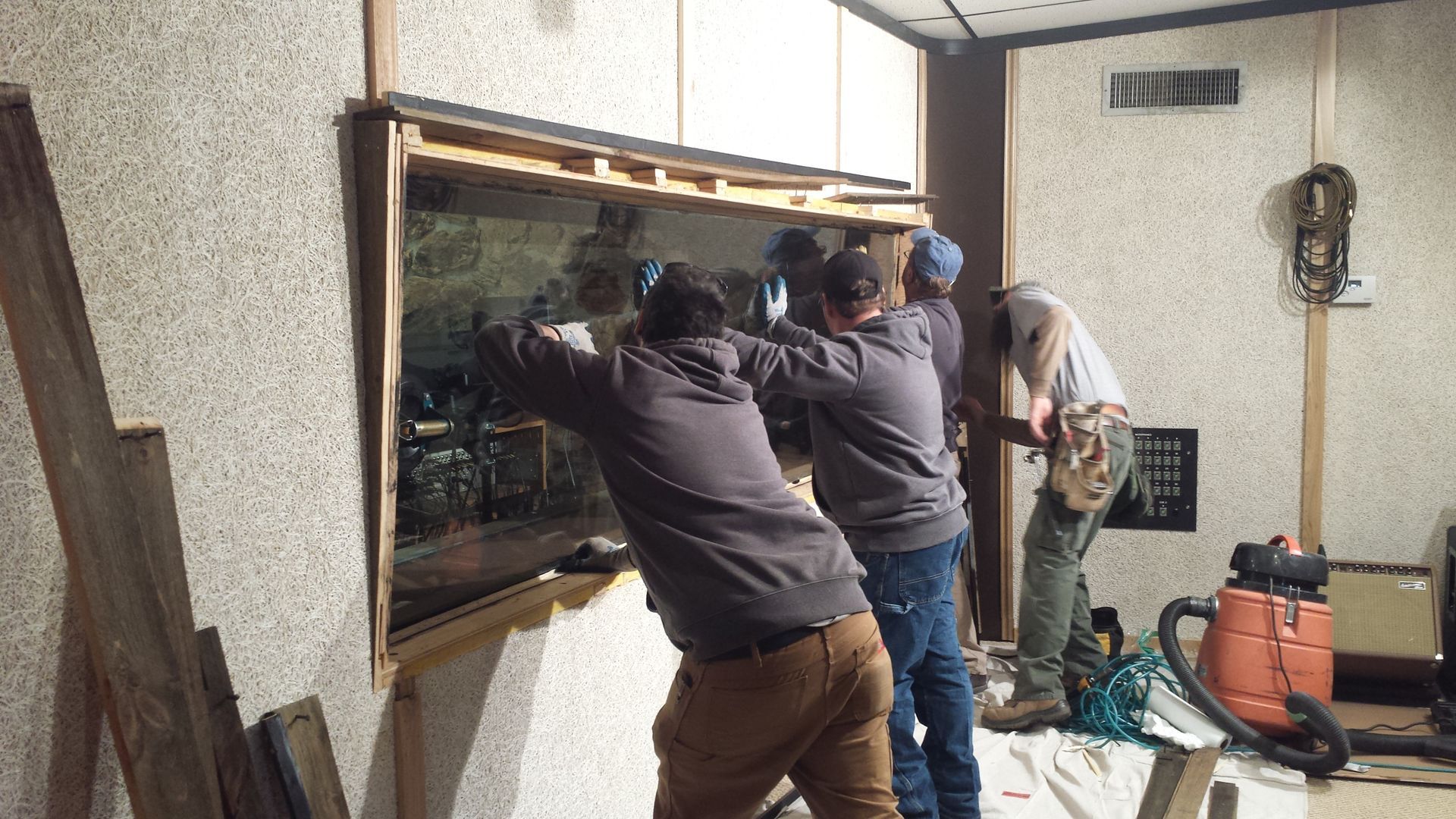 Four people work together to install or remove a large rectangular window pane in a room with textured walls.