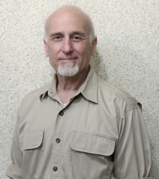 A man with a light goatee, wearing a beige button-down shirt, stands against a textured, off-white wall.