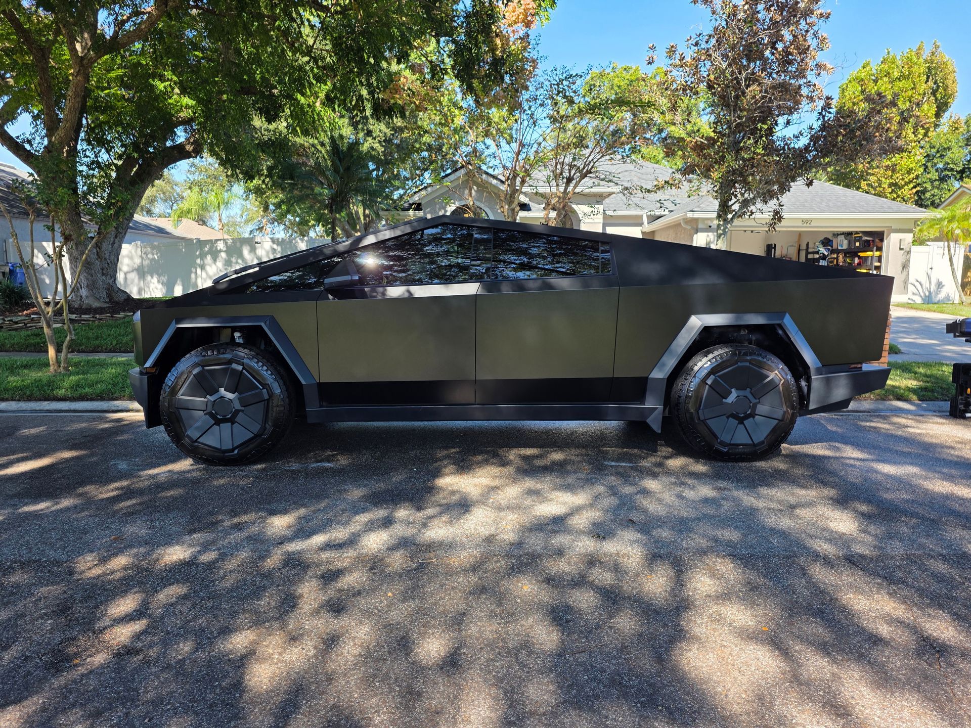 Tesla Cybertruck parked on a paved road, olive green finish with dark wheels, sunny day.
