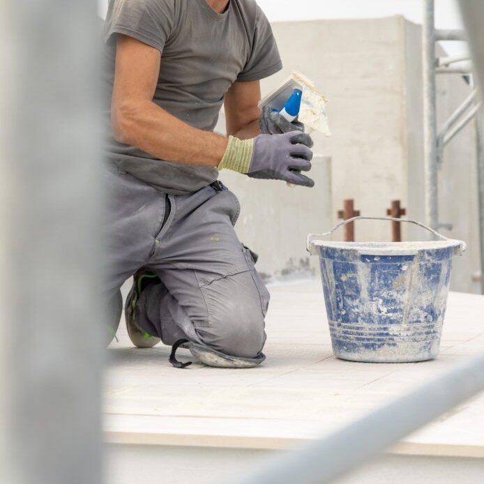 Construction worker kneeling, applying adhesive, next to a bucket. Gray clothing, gloves.